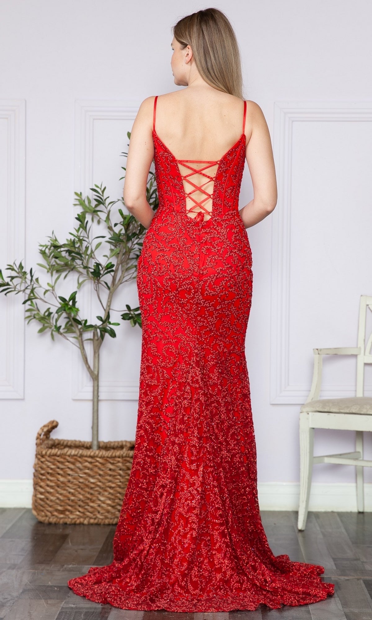 A woman stands facing away in a Glitter-Lace Long Corset Prom Dress 9354, its fitted red silhouette and lace-up back highlighted beside a potted plant and white chair in a softly lit room.